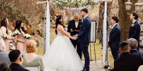 An officiant performing a wedding ceremony for a bride and groom under a floral arch in an outdoor garden setting.