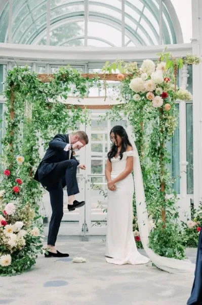 A bride and groom during a wedding ceremony at a floral arch in a glass conservatory.