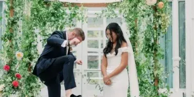 A bride and groom during a wedding ceremony at a floral arch in a glass conservatory.