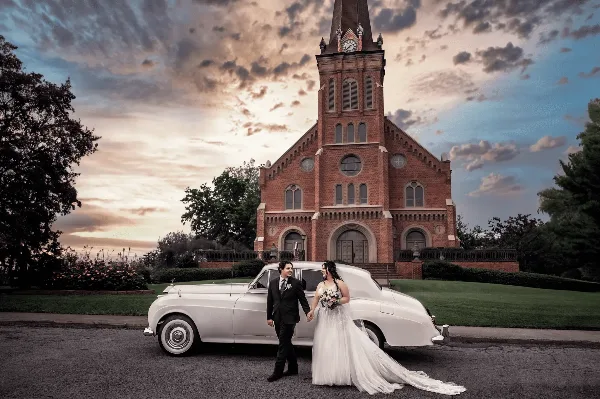 A bride and groom standing next to a white vintage car in front of a brick church at sunset.