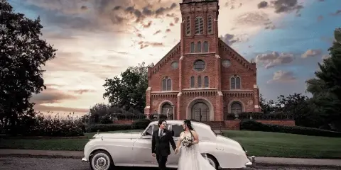 A bride and groom standing next to a white vintage car in front of a brick church at sunset.