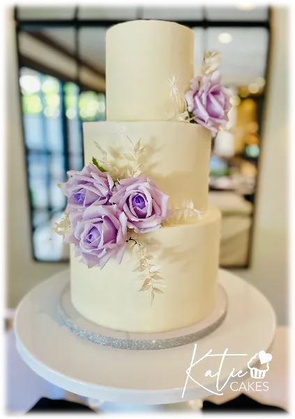 A three-tiered white wedding cake decorated with purple roses and greenery on a white stand.