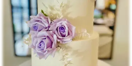 A three-tiered white wedding cake decorated with purple roses and greenery on a white stand.