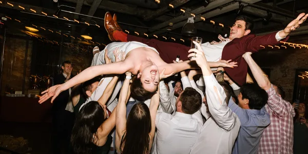 A bride and groom are being tossed into the air by a large, cheering crowd of wedding guests on a dance floor.