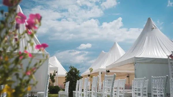 An outdoor event setup featuring white peaked tents and rows of white Chiavari chairs under a blue sky.