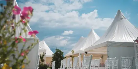 An outdoor event setup featuring white peaked tents and rows of white Chiavari chairs under a blue sky.