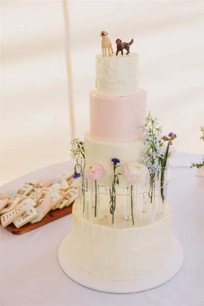 A four-tiered white and pink wedding cake decorated with fresh flowers and dog figurines, displayed next to a tray of custom decorated cookies.
