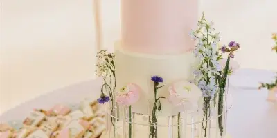 A four-tiered white and pink wedding cake decorated with fresh flowers and dog figurines, displayed next to a tray of custom decorated cookies.