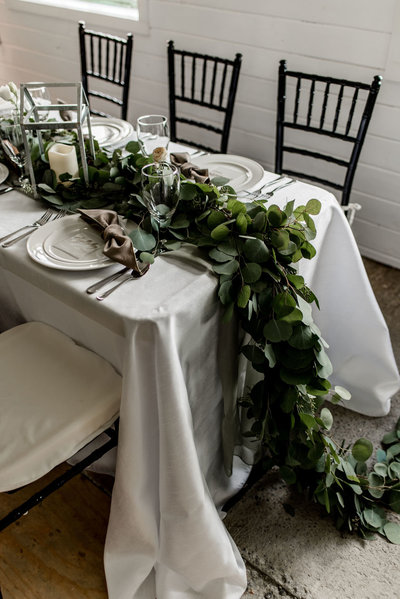 A wedding table setting featuring a white tablecloth, black chiavari chairs, and a long eucalyptus greenery runner.