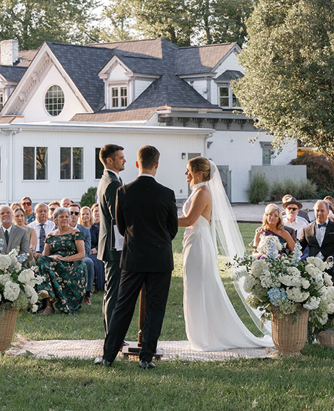 A bride and groom exchanging vows during an outdoor wedding ceremony in front of a historic white mansion.