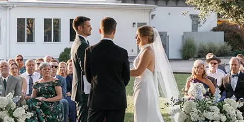 A bride and groom exchanging vows during an outdoor wedding ceremony in front of a historic white mansion.