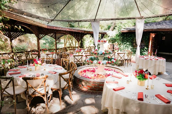 An outdoor garden event venue featuring round tables with white tablecloths, red napkins, and a central floral centerpiece under a canopy.