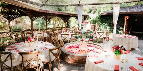 An outdoor garden event venue featuring round tables with white tablecloths, red napkins, and a central floral centerpiece under a canopy.
