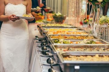 A bride in a white wedding dress serving food from a buffet line of chafing dishes.
