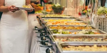 A bride in a white wedding dress serving food from a buffet line of chafing dishes.