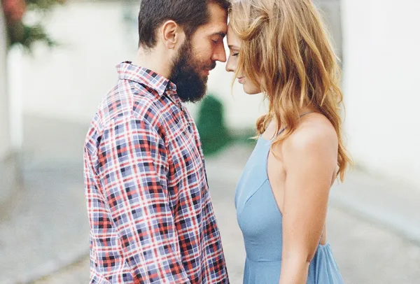 A bearded man in a plaid shirt and a woman in a blue dress standing close together with their foreheads touching.