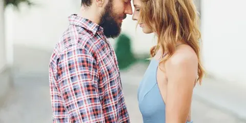 A bearded man in a plaid shirt and a woman in a blue dress standing close together with their foreheads touching.