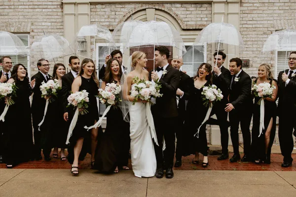 A wedding party including the bride and groom and their attendants standing outdoors with clear umbrellas.