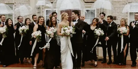 A wedding party including the bride and groom and their attendants standing outdoors with clear umbrellas.