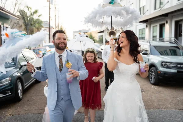 A bride and groom walking down a street holding feathered umbrellas, with a bridesmaid following behind.