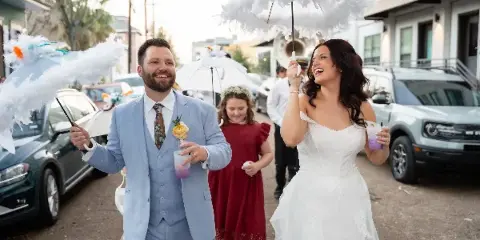A bride and groom walking down a street holding feathered umbrellas, with a bridesmaid following behind.