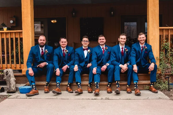 A groom and five groomsmen wearing matching blue suits and red ties sitting on a wooden porch.