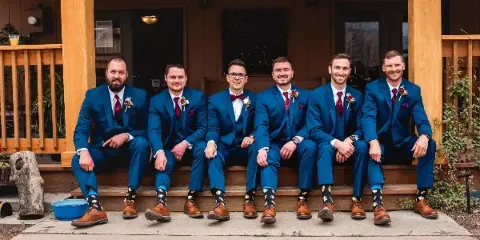 A groom and five groomsmen wearing matching blue suits and red ties sitting on a wooden porch.