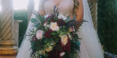 A bride in a white wedding dress holding a large, cascading bouquet of white and burgundy flowers with greenery.