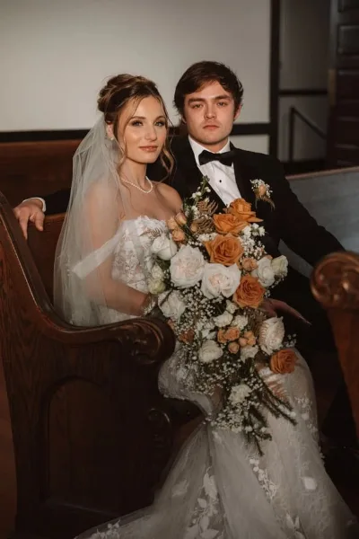 A bride and groom posing together, with the bride holding a large bouquet of white and orange roses and baby's breath.
