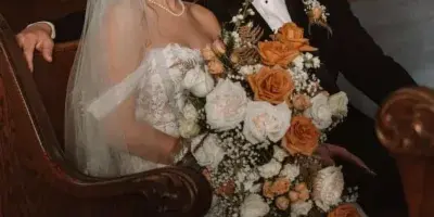 A bride and groom posing together, with the bride holding a large bouquet of white and orange roses and baby's breath.