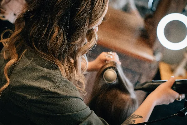 A stylist blow-drying and styling a client's hair with a round brush in a salon.