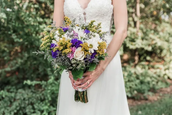 A bride in a white lace dress holding a colorful bouquet of purple, white, and yellow flowers with greenery.