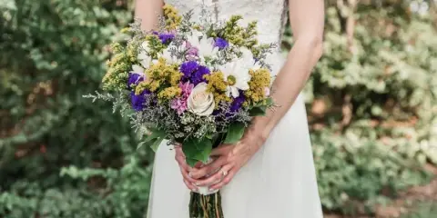 A bride in a white lace dress holding a colorful bouquet of purple, white, and yellow flowers with greenery.