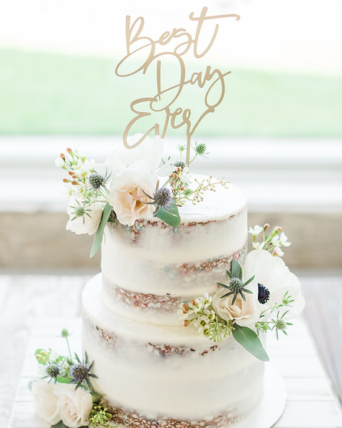 A three-tier semi-naked wedding cake decorated with white and blush flowers, greenery, and a gold 'Best Day Ever' topper.