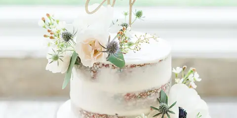 A three-tier semi-naked wedding cake decorated with white and blush flowers, greenery, and a gold 'Best Day Ever' topper.
