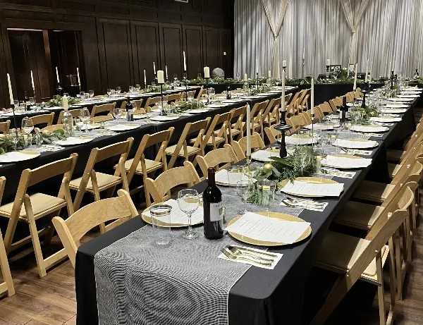 A large event venue featuring long banquet tables set with black tablecloths, gold-rimmed plates, white candles, and wooden chairs.