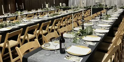 A large event venue featuring long banquet tables set with black tablecloths, gold-rimmed plates, white candles, and wooden chairs.