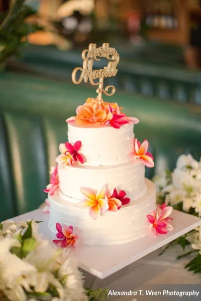 Three-tier white wedding cake decorated with pink and yellow plumeria flowers and a gold 'Aloha Maui' topper, displayed on a white cake stand.