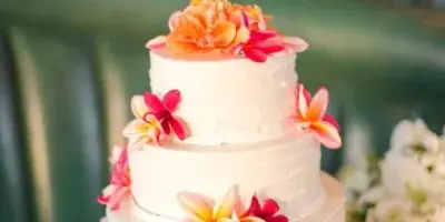 Three-tier white wedding cake decorated with pink and yellow plumeria flowers and a gold 'Aloha Maui' topper, displayed on a white cake stand.
