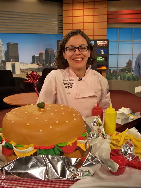A baker in a white chef's coat embroidered with 'Adrienne & Company Bakery Cafe' stands behind a large, custom cake shaped like a hamburger with fondant toppings.