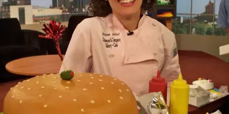 A baker in a white chef's coat embroidered with 'Adrienne & Company Bakery Cafe' stands behind a large, custom cake shaped like a hamburger with fondant toppings.