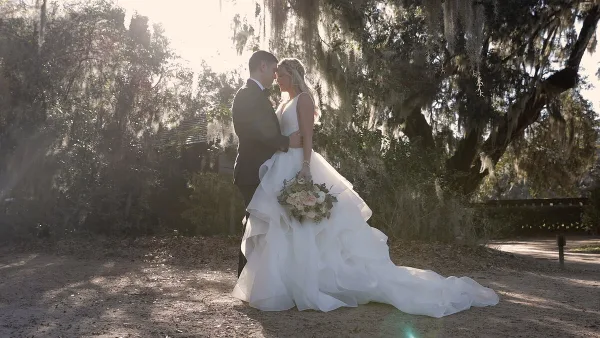A bride and groom embracing in a sunlit outdoor setting with Spanish moss hanging from trees.