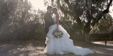 A bride and groom embracing in a sunlit outdoor setting with Spanish moss hanging from trees.