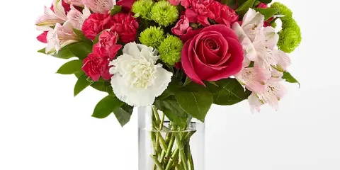 A vibrant floral bouquet featuring pink roses, white carnations, and green chrysanthemums arranged in a clear cylindrical glass vase.