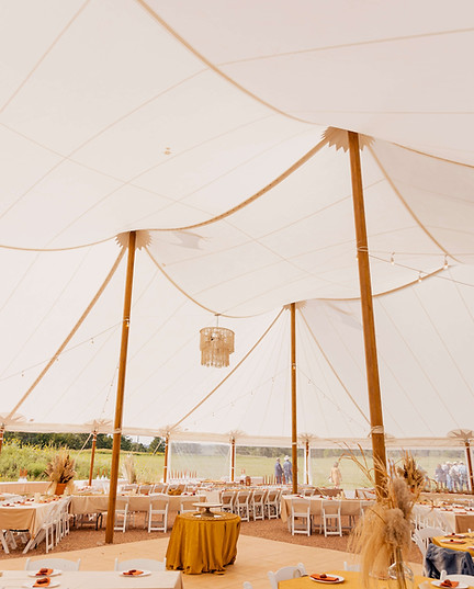 Interior view of a large white wedding tent with wooden support poles, set up with round tables, white chairs, and a hanging chandelier.