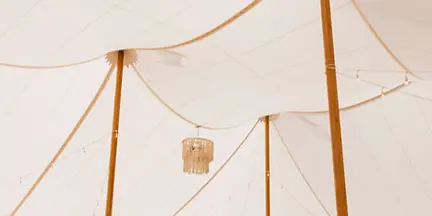 Interior view of a large white wedding tent with wooden support poles, set up with round tables, white chairs, and a hanging chandelier.