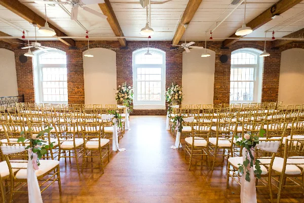 Interior view of a wedding venue with exposed brick walls, high ceilings, and rows of gold chairs arranged for a ceremony.