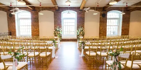Interior view of a wedding venue with exposed brick walls, high ceilings, and rows of gold chairs arranged for a ceremony.