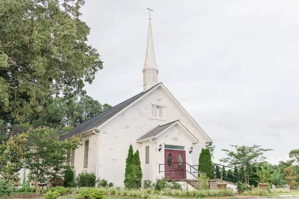 Exterior view of a white wooden chapel with a tall steeple and red double doors, surrounded by green landscaping.