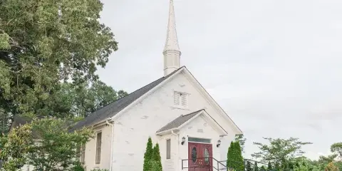 Exterior view of a white wooden chapel with a tall steeple and red double doors, surrounded by green landscaping.